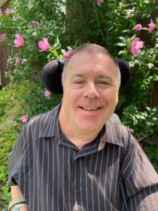 Male seated in chair, smiling with a black and blue striped shirt. Pink flowers in the background.