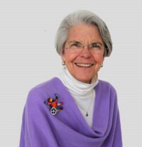 Photo of a woman smiling with excitement; she wearing glasses, a white turtle neck and a purple poncho. On the poncho is a pin of a person exhibiting joy using a wheelchair.