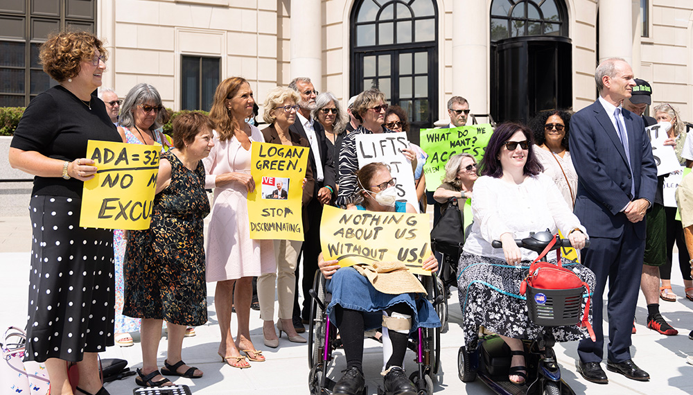 A group of individuals crowd around the court house. Among these people a some individuals with very apparent mobility disabilities. All are holding up signs advocating for the viability and right to use Lyft services.